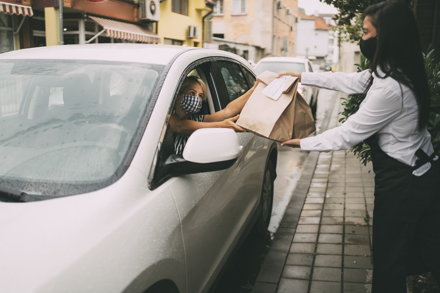 Waitress wearing protective face mask is giving disposable package with food  to pretty smiling female driver. Reopening after COVID-19 concepts.