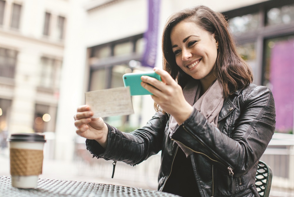 A beautiful Caucasian adult woman sits in a New York city park, taking a picture of a check with her smart phone for a  Remote Deposit Capture.  She smiles, wearing modern stylish clothing with darker and black colors.