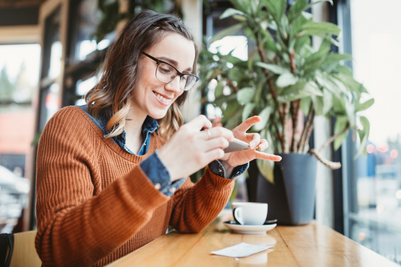 A smiling young woman takes a picture with her smart phone of a check or paycheck for digital electronic depositing, also known as "Remote Deposit Capture".  She sits in a coffee shop, enjoying an espresso latte.  Bright sunlight shines in the window.  Horizontal image.