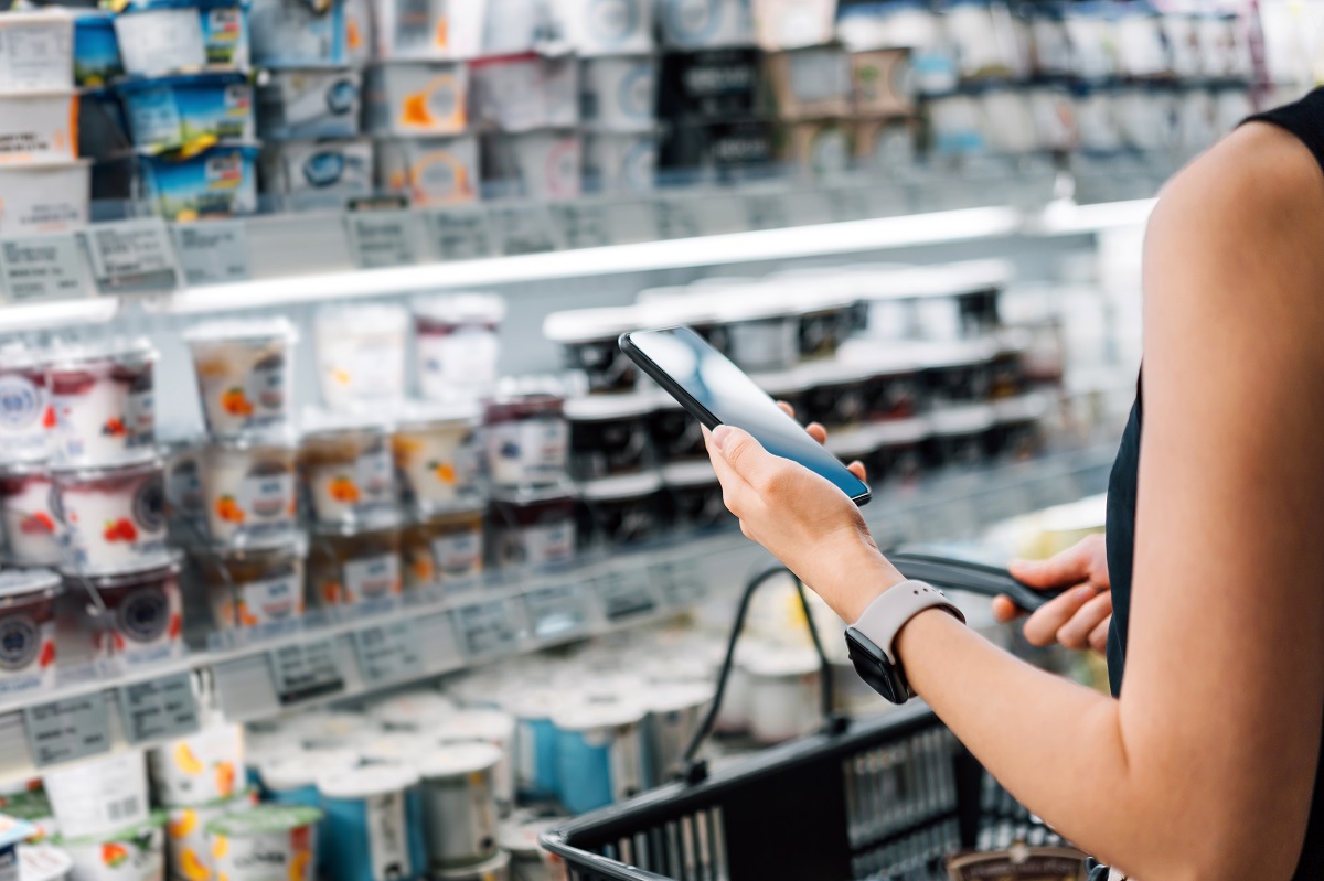 Cropped shot of young Asian woman holding shopping basket and checking her shopping list on smartphone while grocery shopping in a supermarket
