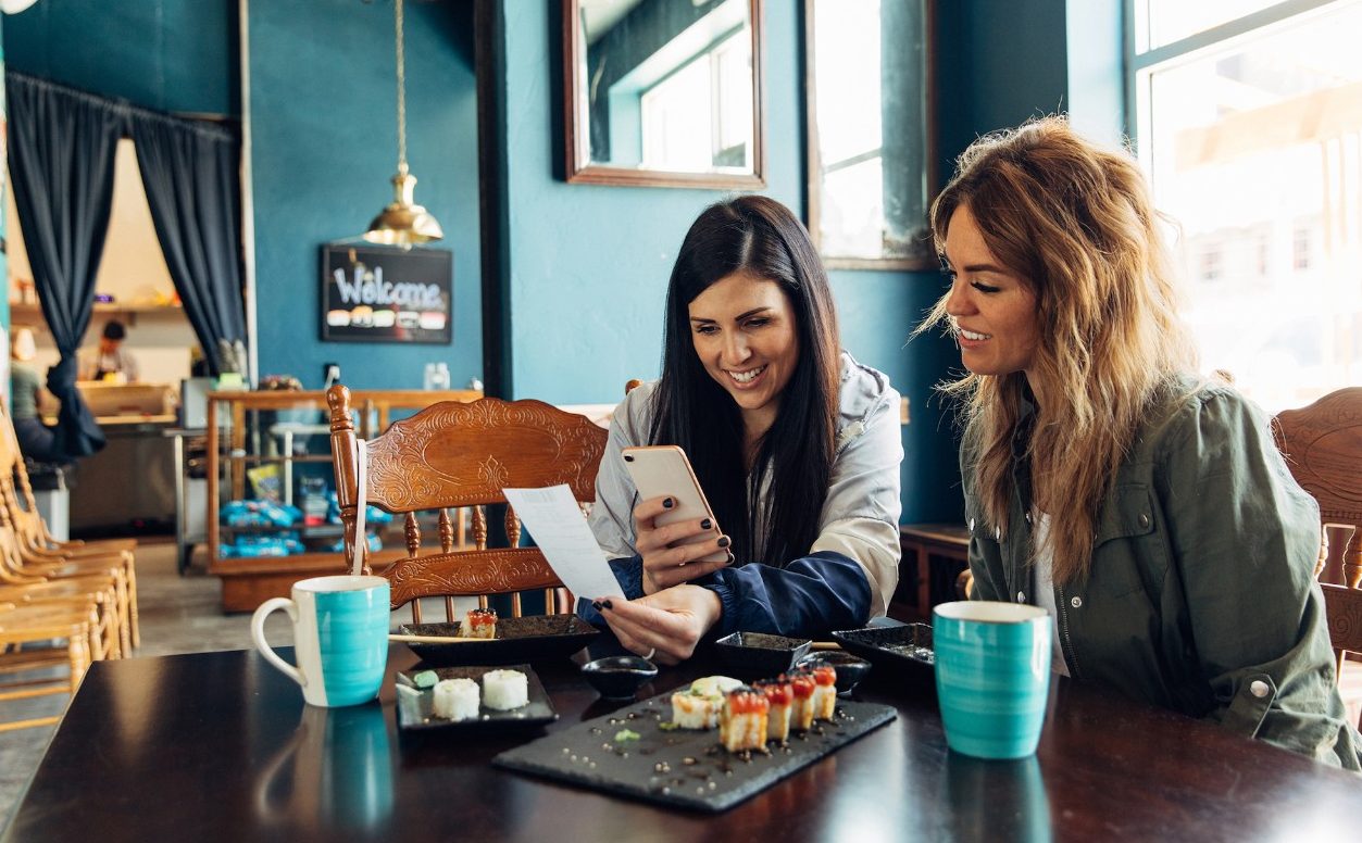Two women of the Millennial Generation are taking a picture of a restaurant receipt after eating lunch at a local sushi restaurant. The woman is using her bank's app to balance her monthly budget. Image taken in Utah, USA.